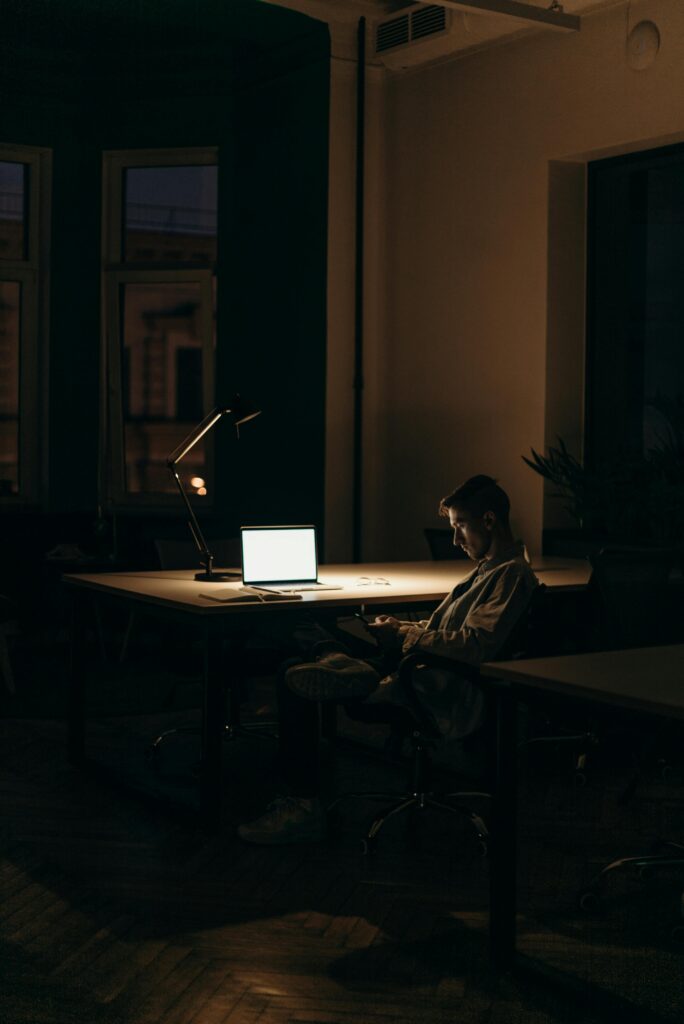 A lone man working on a laptop under dim lighting in a quiet office at night.